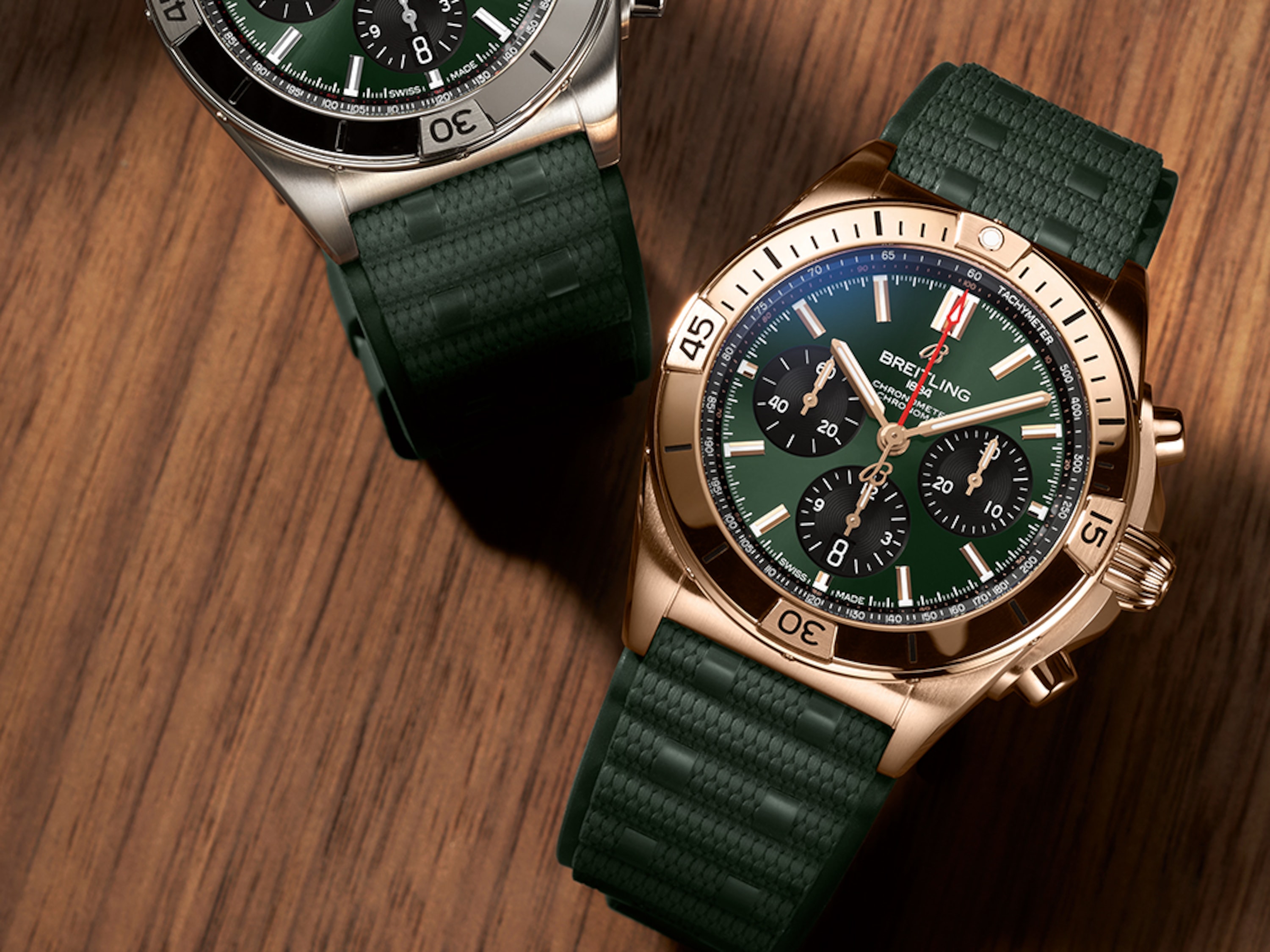 Un Chronomat de oro y otro de plata con correas de caucho verde oscuro sobre una mesa marrón.