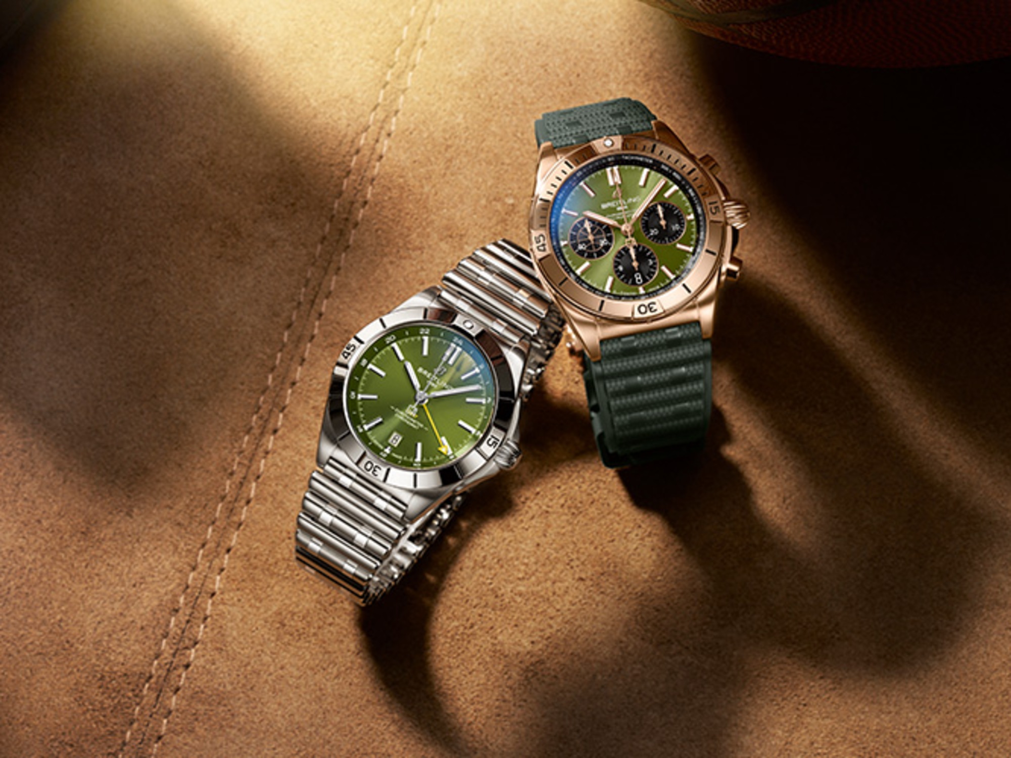 A gold and a silver Chronomat with dark green rubber straps on a brown table.
