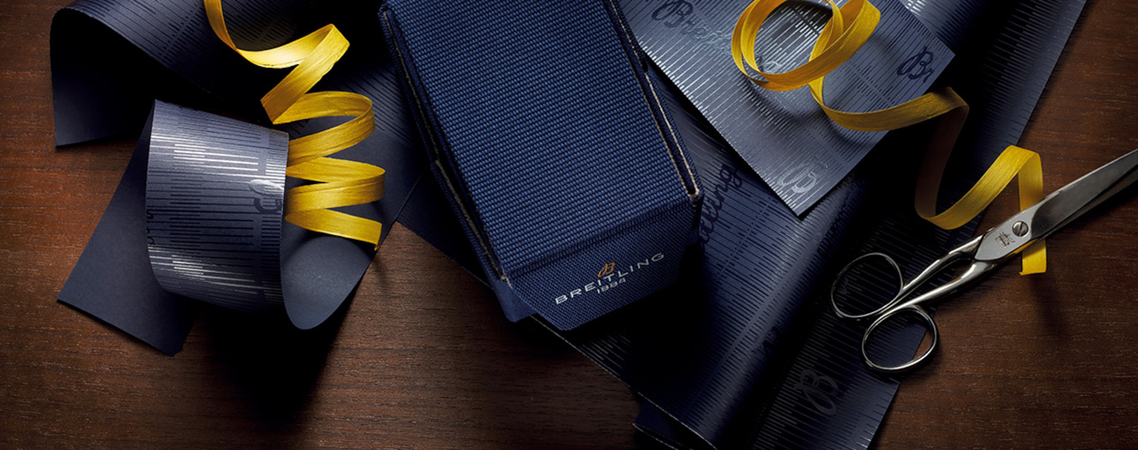 {generated} Breitling gift packaging with a navy box, branded wrap and golden ribbon arranged neatly on a wooden surface