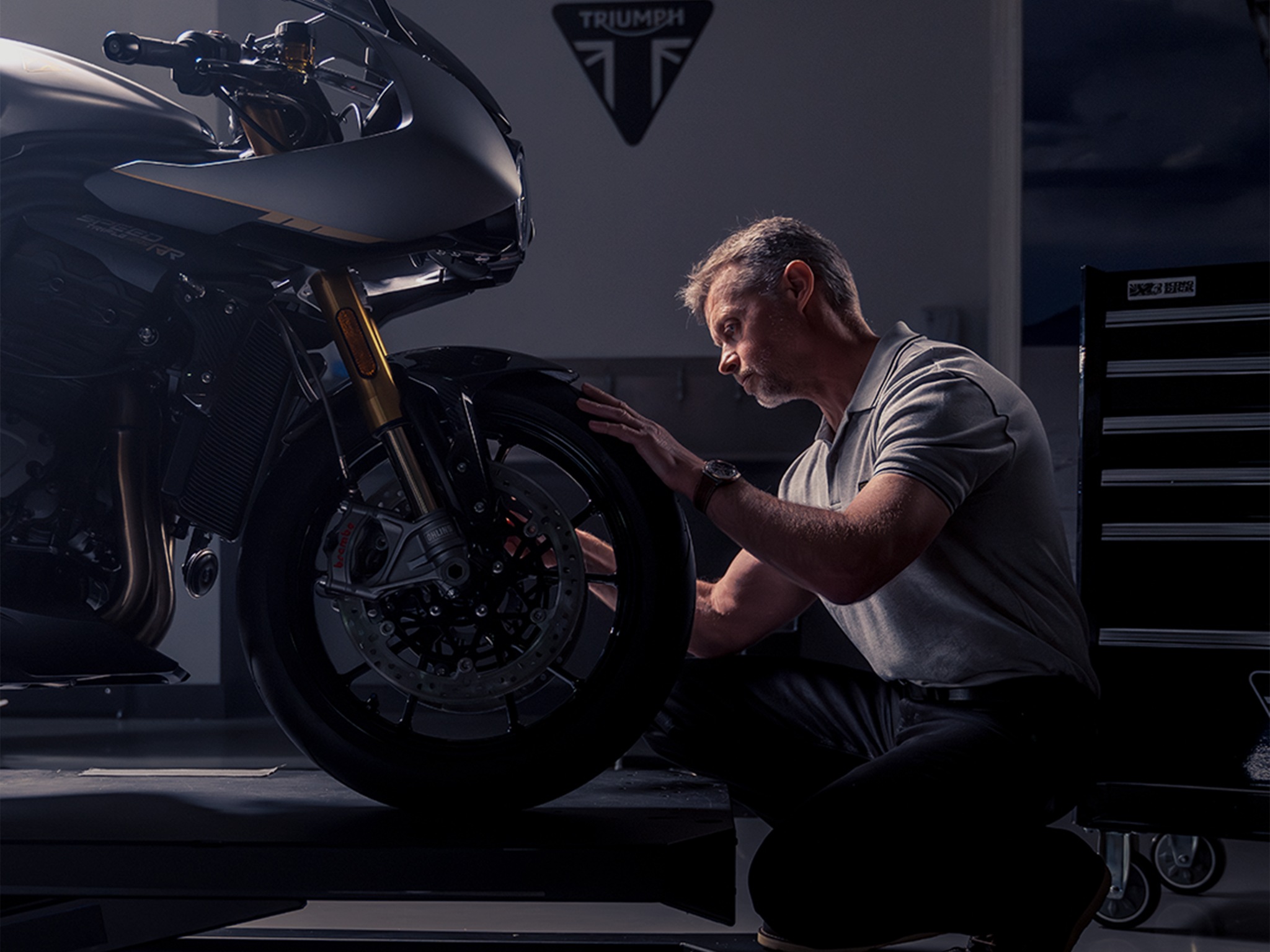 Un hombre con un reloj Breitling en el punto de mira se arrodilla ante una moto Triumph