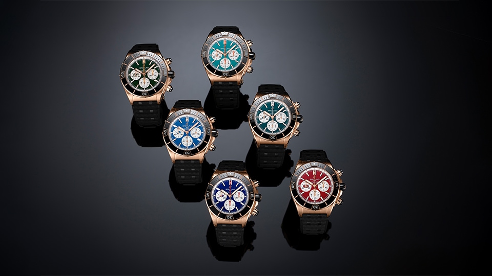 Six Breitling chronograph watches with gold cases and black rubber straps, featuring colorful dials in green, blue, and red against a dark background.