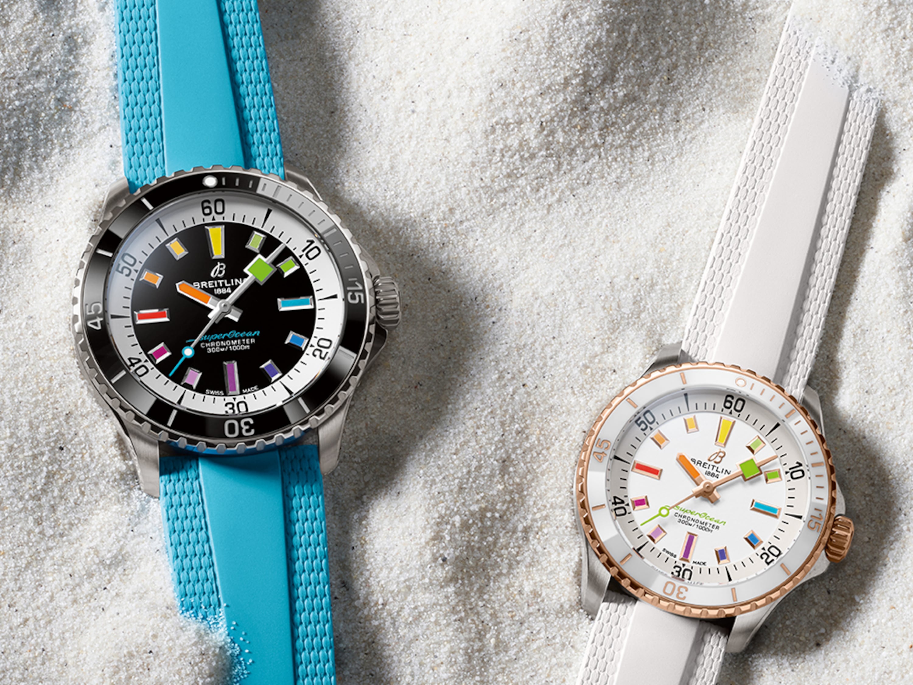 Two Superocean Automatic watches, one with a turquoise strap and one with a white strap, lie in the sand.