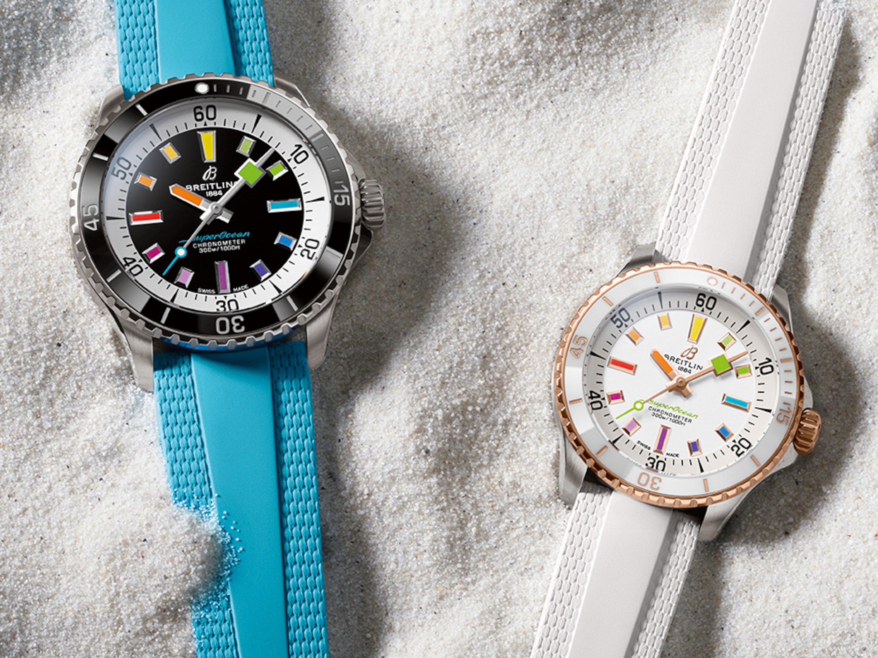 Two Superocean Automatic watches, one with a turquoise strap and one with a white strap, lie in the sand.
