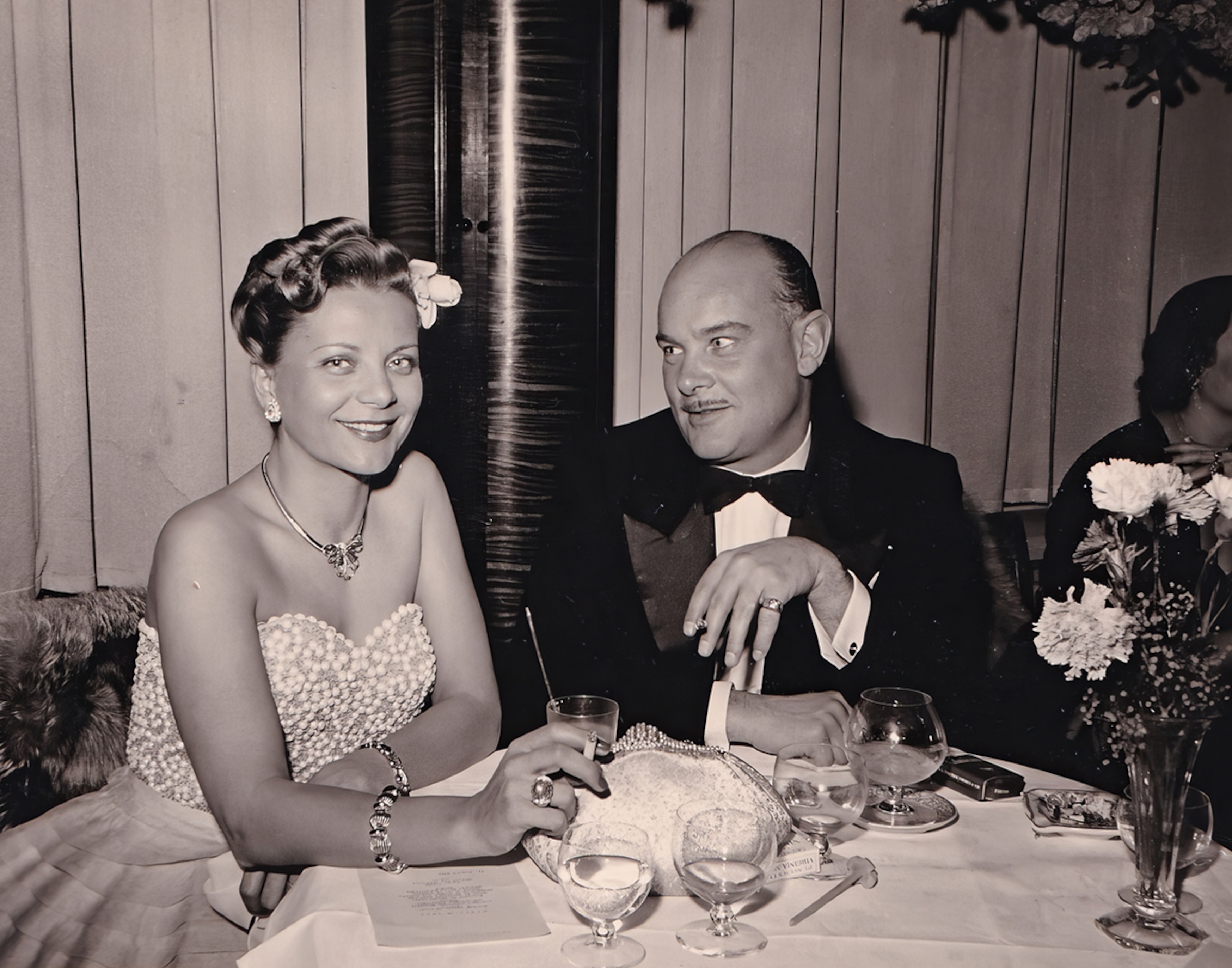 {generated} Elegante Frau in Vintage-Abendgarderobe sitzt an einem formellen Tisch neben einem Mann und vermittelt zeitlosen Stil und Anmut.