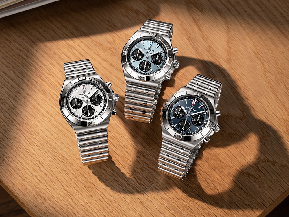 A gold and a silver Chronomat with dark green rubber straps on a brown table.