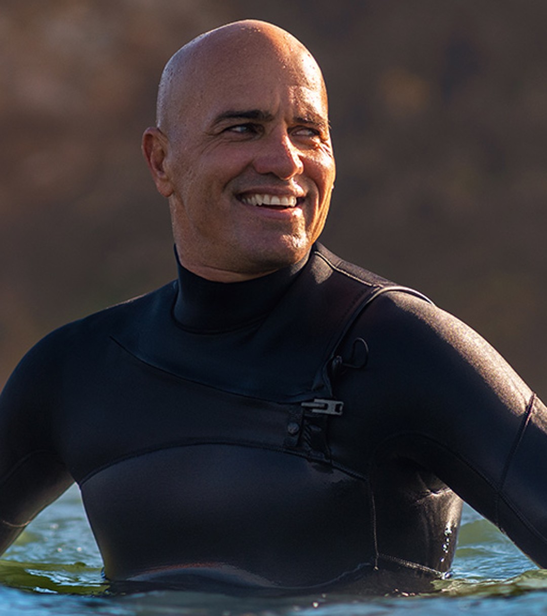 {generated} Un surfeur en combinaison se tient dans une eau côtière calme, la lumière du soleil illuminant les vagues devant une falaise floue