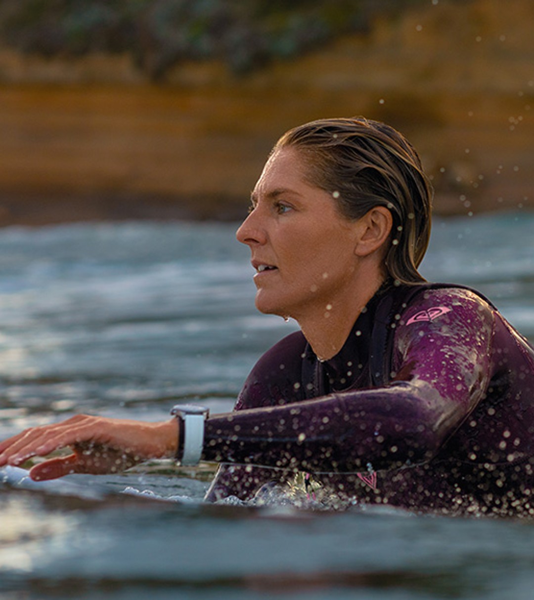 {generated} A woman paddling a surfboard at sunset, wearing a wetsuit and a Breitling watch, with coastal cliffs in the background.