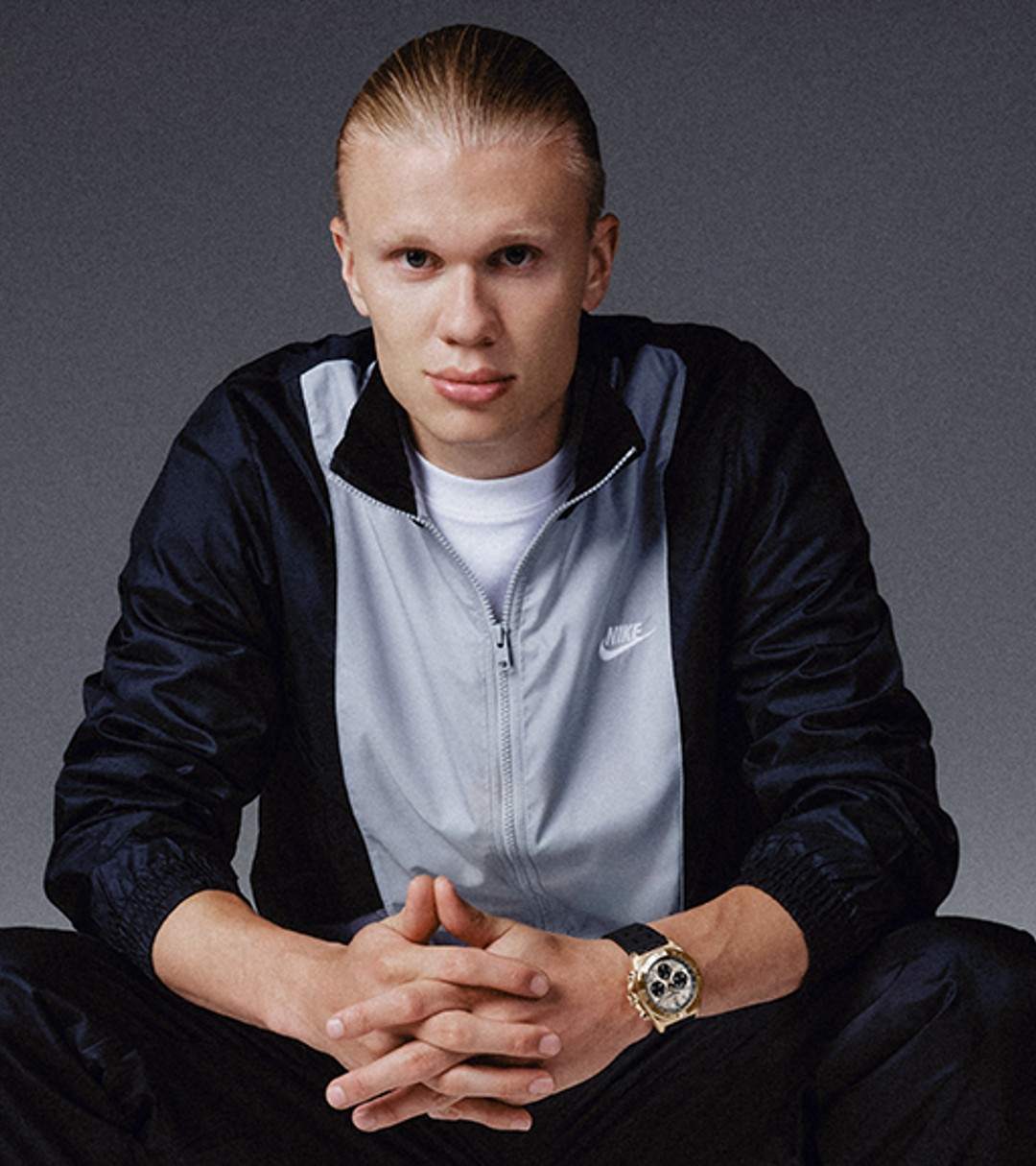 {generated} A seated male ambassador in sportswear highlights a gold Breitling watch against a dark backdrop with a composed posture.
