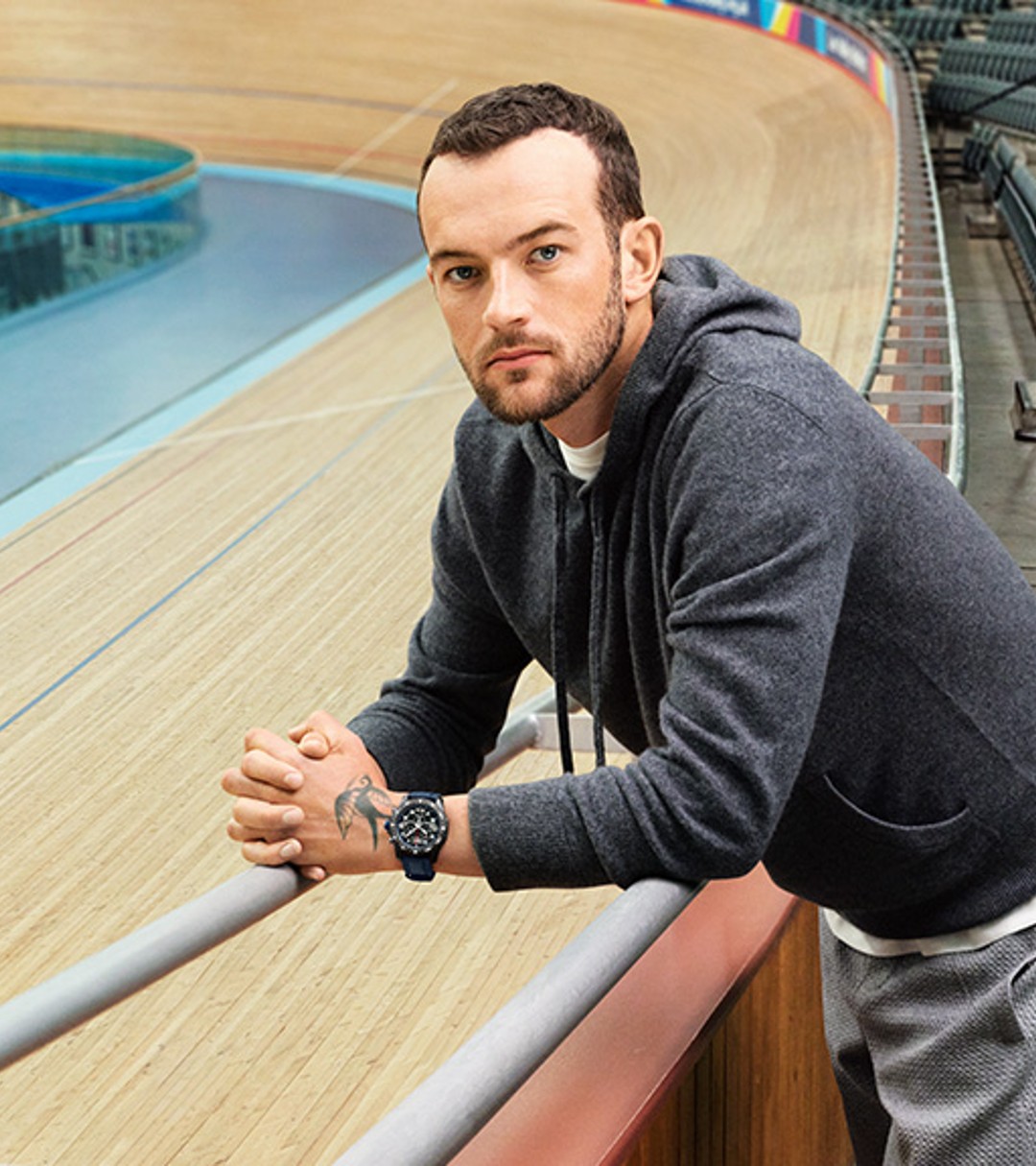 {generated} Man leaning on a velodrome railing wearing a Breitling sports watch with blue strap beside empty tiered seating.