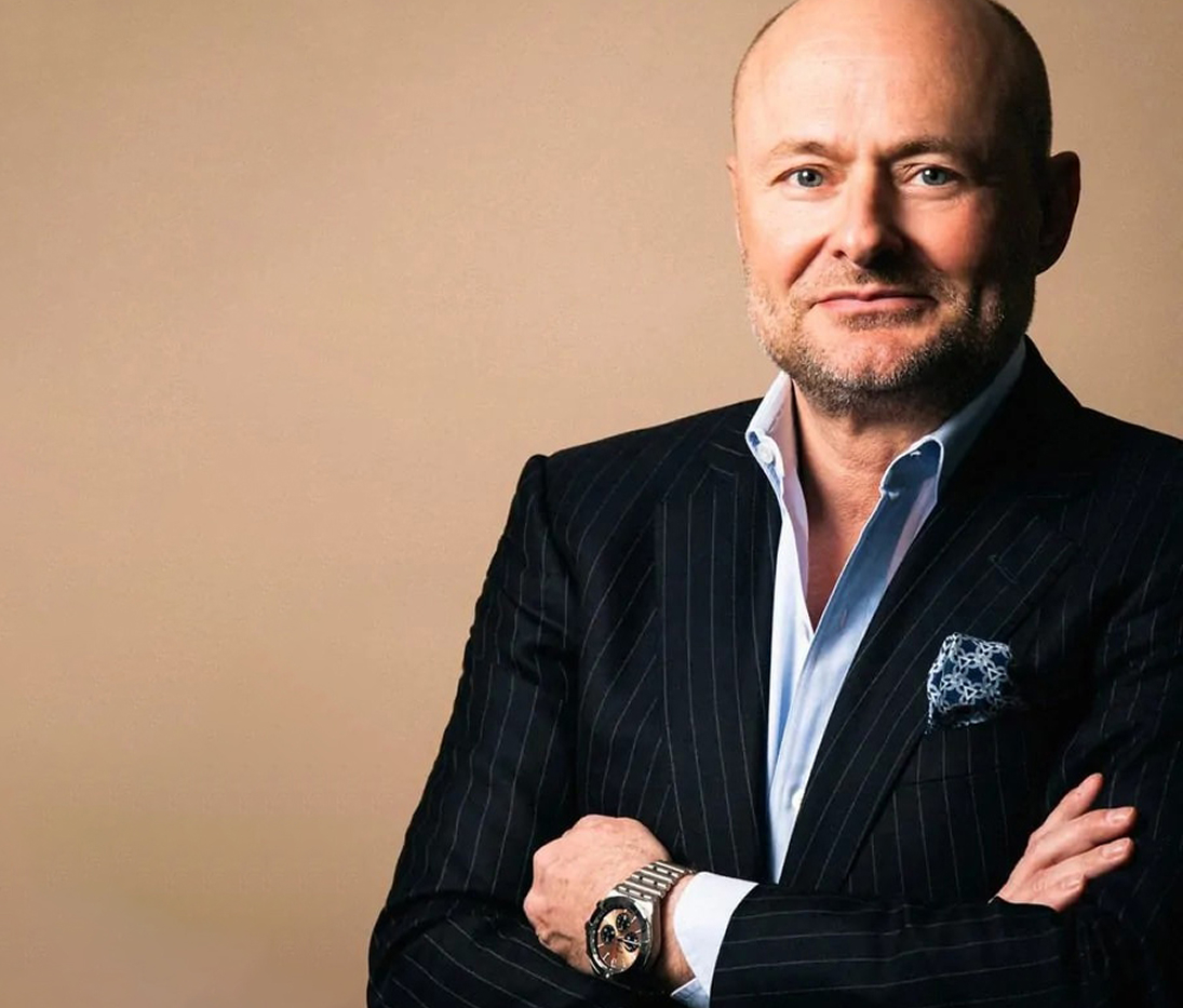 {generated} Un uomo in abito elegante a braccia incrociate mostra un orologio Breitling con bracciale metallico su sfondo caldo e sobrio.