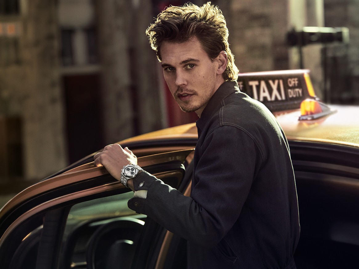 Person in black leather jacket wearing a Breitling watch, standing next to motorcycle on outdoor film set with lighting equipment.