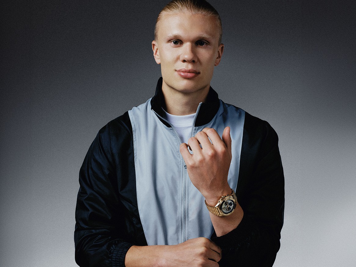 {generated} A man in sporty jacket raising his hand, showcasing a Breitling watch on his wrist against a sleek gradient backdrop.