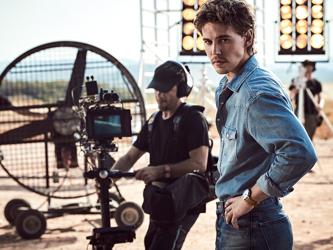 {generated} A male ambassador on a film set wearing a Breitling watch, standing near crew and lighting equipment in a sunlit outdoor scene.
