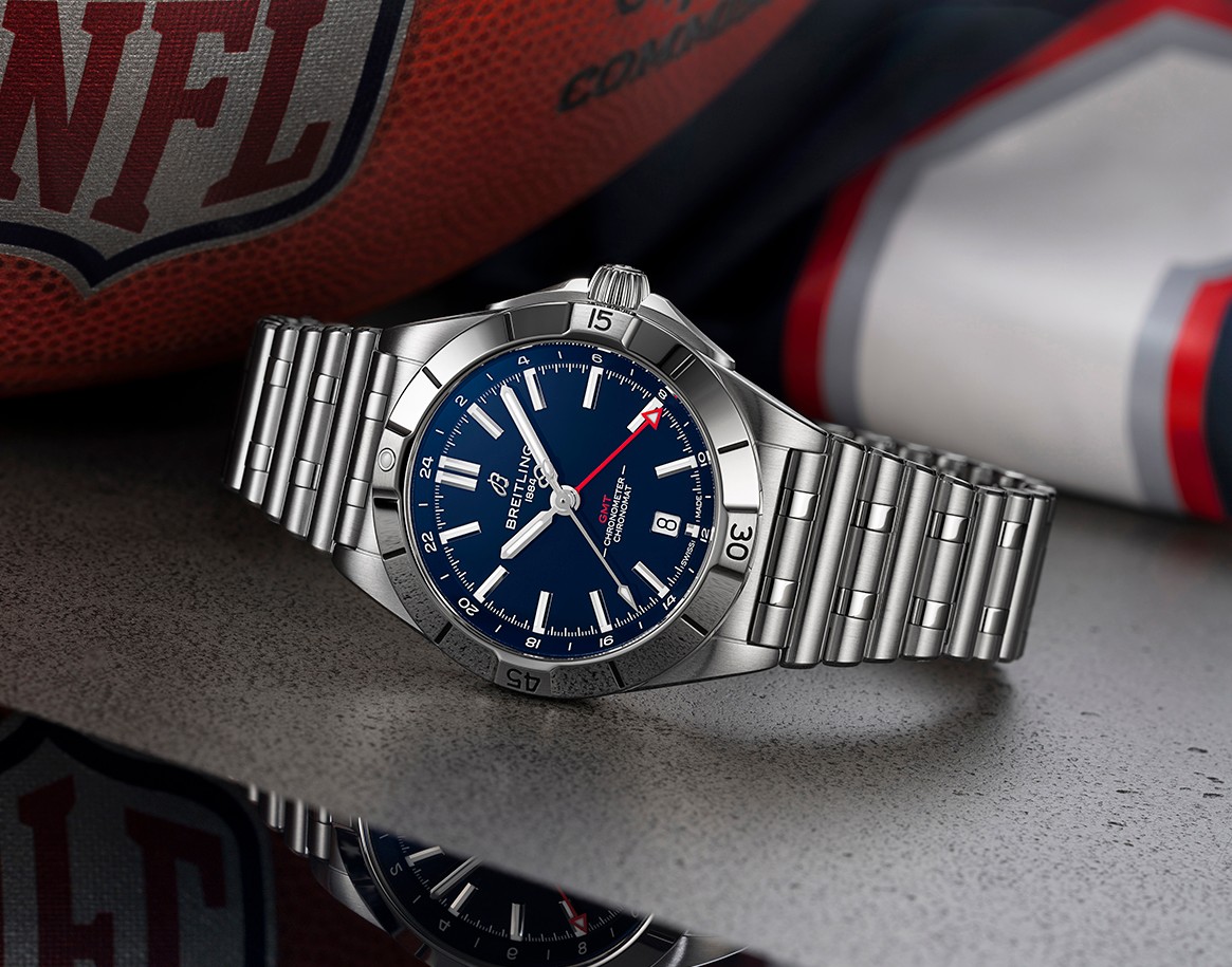 {generated} Breitling Chronomat GMT com mostrador azul e bracelete de aço ao lado de itens NFL nas cores do New England Patriots.