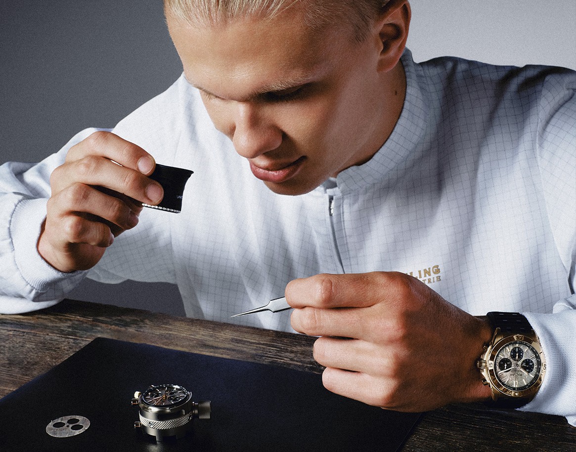 {generated} Man inspecting watch components with precision tools, wearing a gold Breitling chronograph and branded Breitling attire.