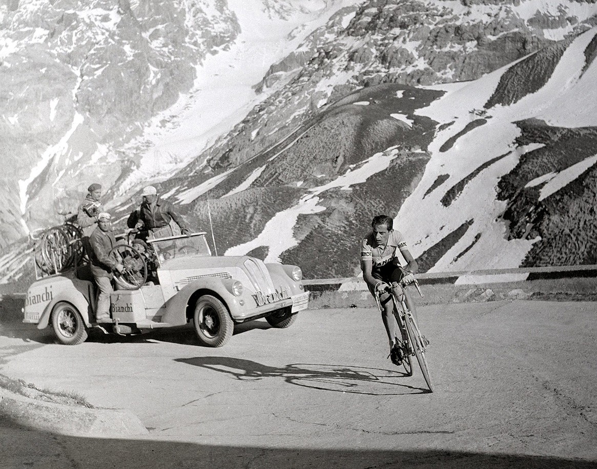 {generated} Cena em estrada montanhosa com ciclista à frente de carro de apoio junto a picos nevados, capturando um momento histórico.
