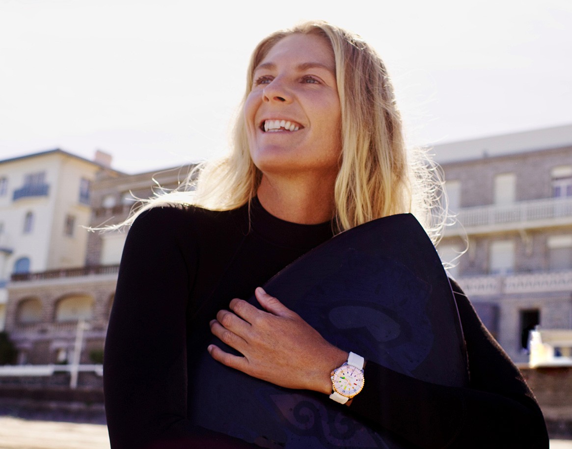 {generated} Stephanie Gilmore holding a surfboard on a sunny beach, wearing a white‑strap Breitling watch shown prominently.