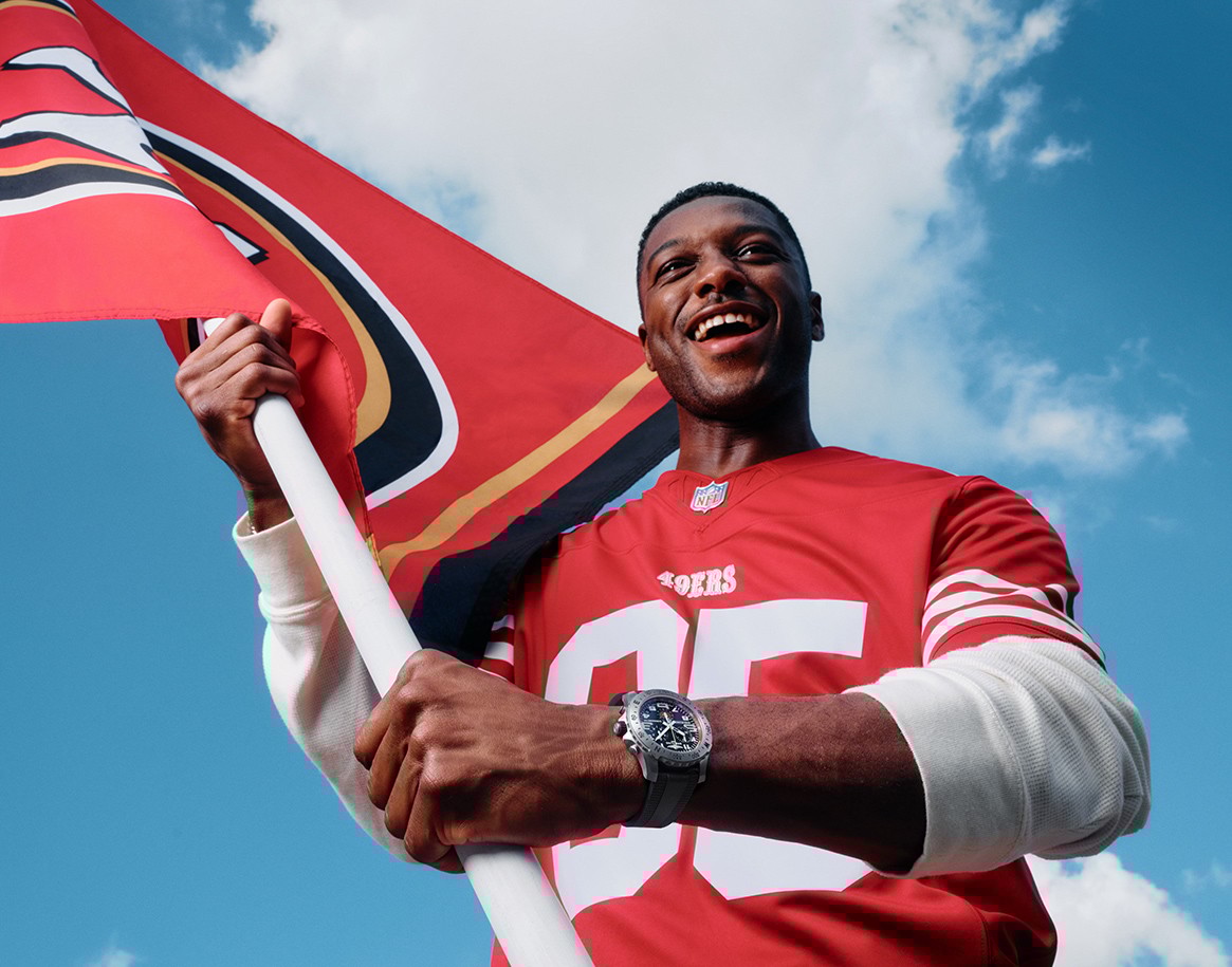 {generated} 穿紅色NFL球衣的球迷高舉球隊旗幟，在明亮天空下展示百年靈NFL球隊特別版腕錶。