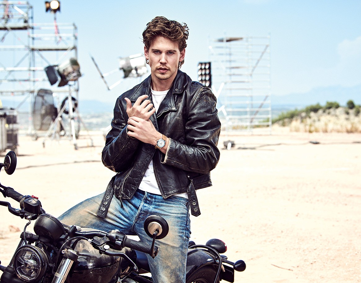 {generated} Austin seated on a motorbike in a desert set, wearing a leather jacket and highlighting a Breitling watch on his wrist.
