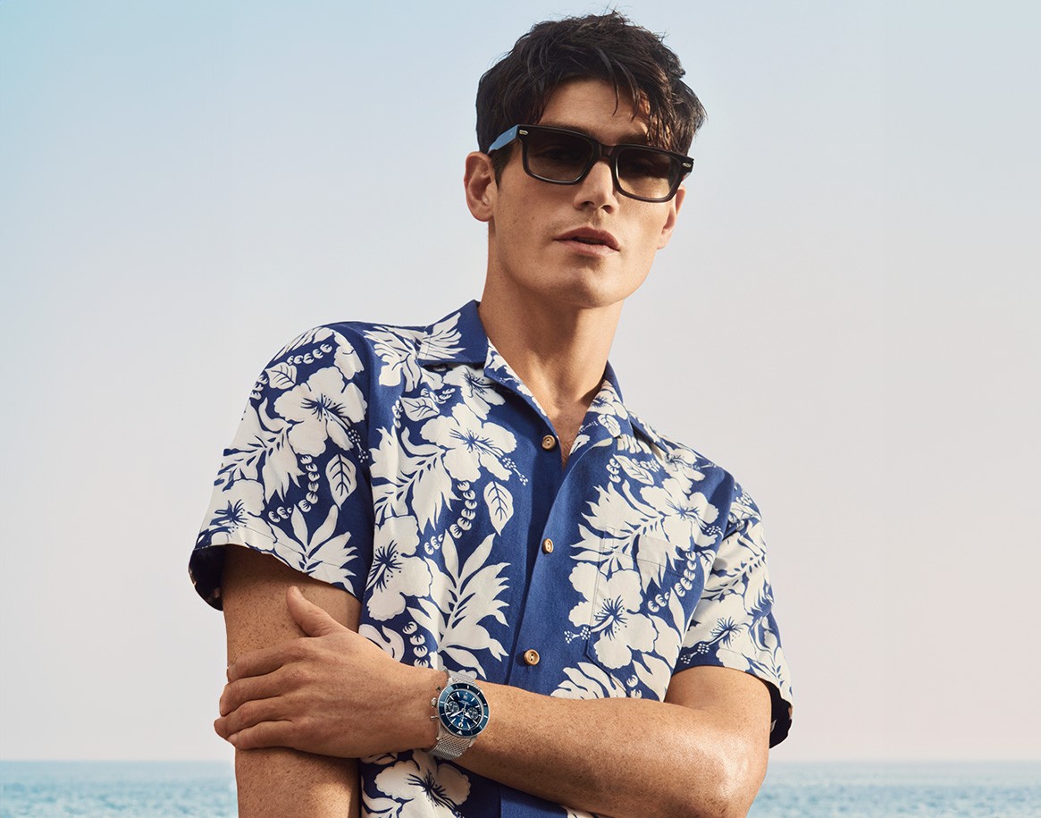 {generated} Man by the sea wearing sunglasses and a floral shirt, showcasing a Breitling watch in a relaxed coastal style.