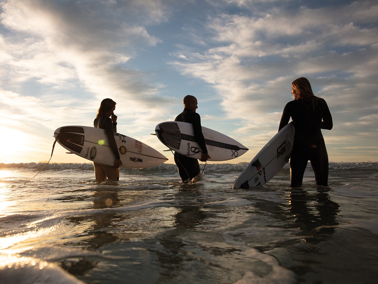 {generated} Tre surfisti avanzano nel mare al tramonto con le loro tavole, mentre le onde si alzano intorno creando un’atmosfera dinamica.