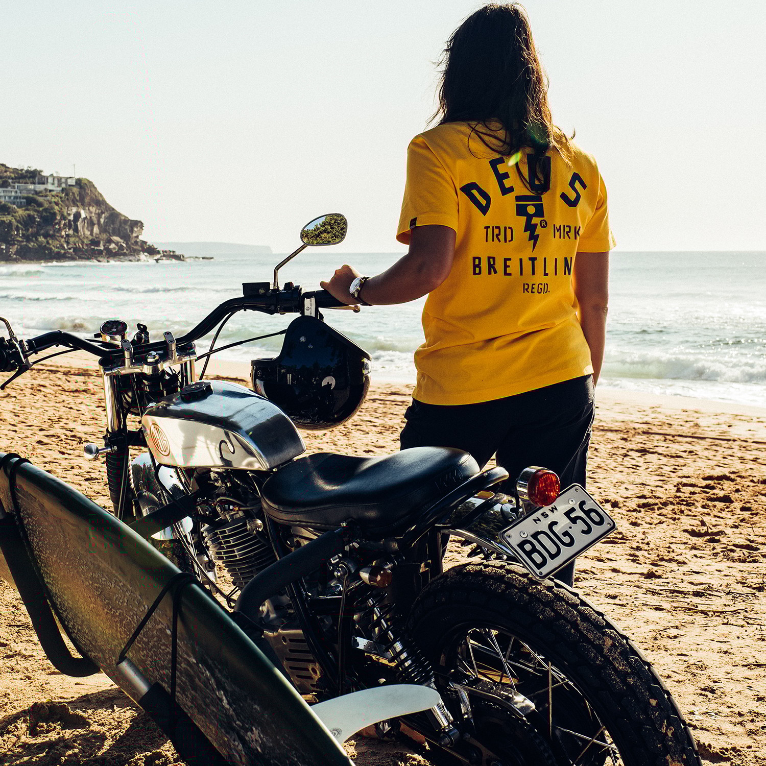 {generated} A person in a yellow Deus x Breitling shirt stands beside a surfboard‑equipped motorcycle on a sunny beach.