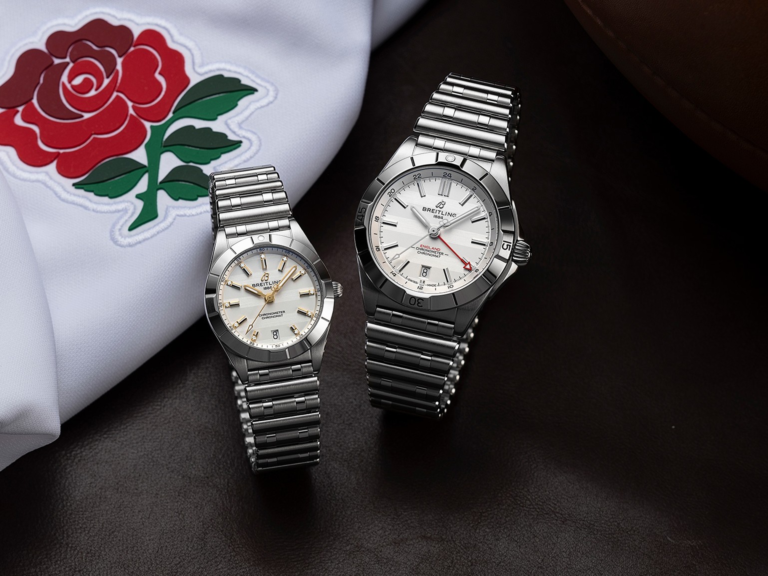 {generated} 兩款Breitling Chronomat England腕錶陳列在英格蘭玫瑰徽章旁，以細膩拋光的鋼質外觀展現典雅氣度，並突顯其精密工藝與運動格調，塑造出獨特英倫風範與卓越品質感，契合六國橄欖球賽精神與品牌傳承。