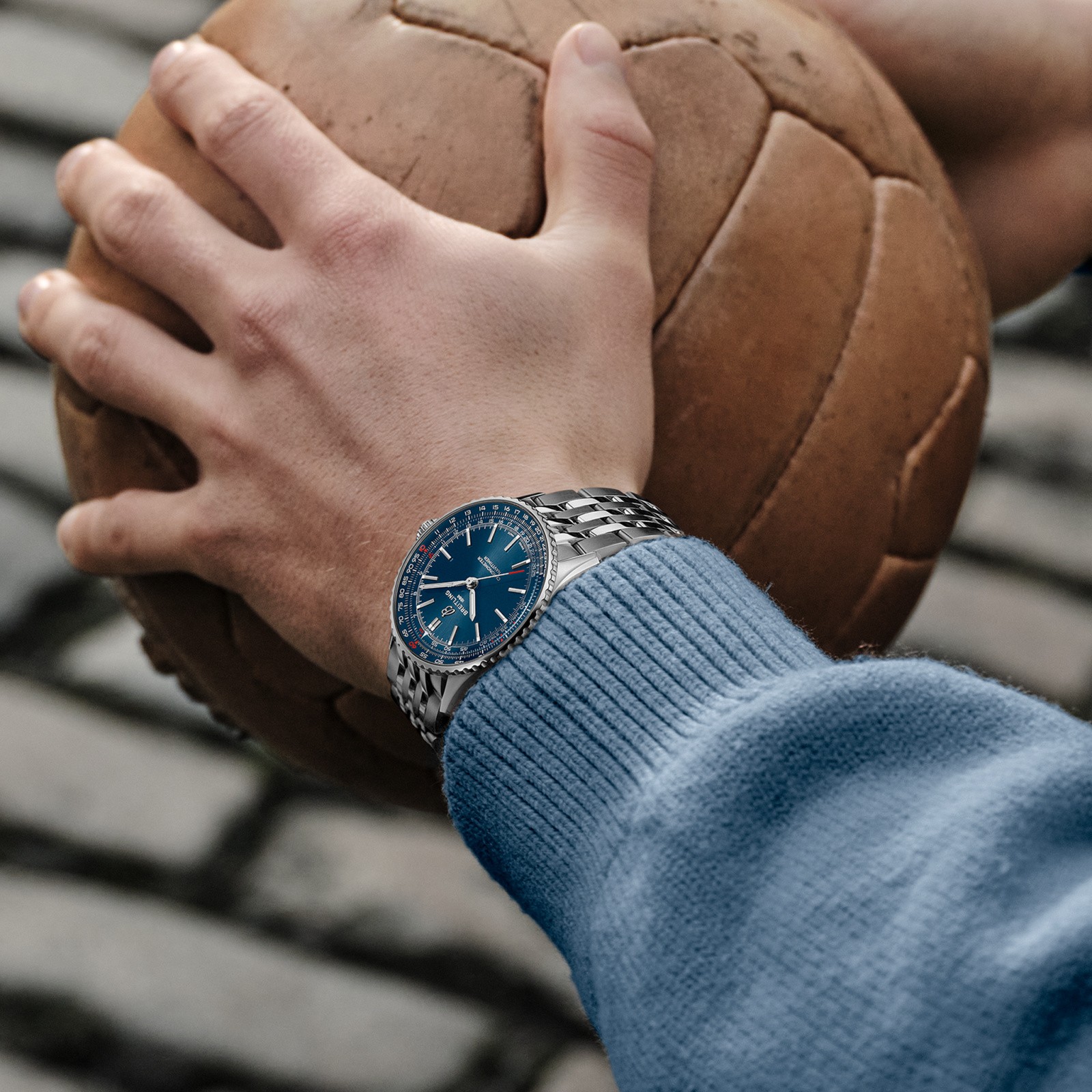 {generated} 手握復古皮球，腕上佩戴藍色錶盤與鋼錶鏈的Breitling Navitimer，展現戶外休閒質感。