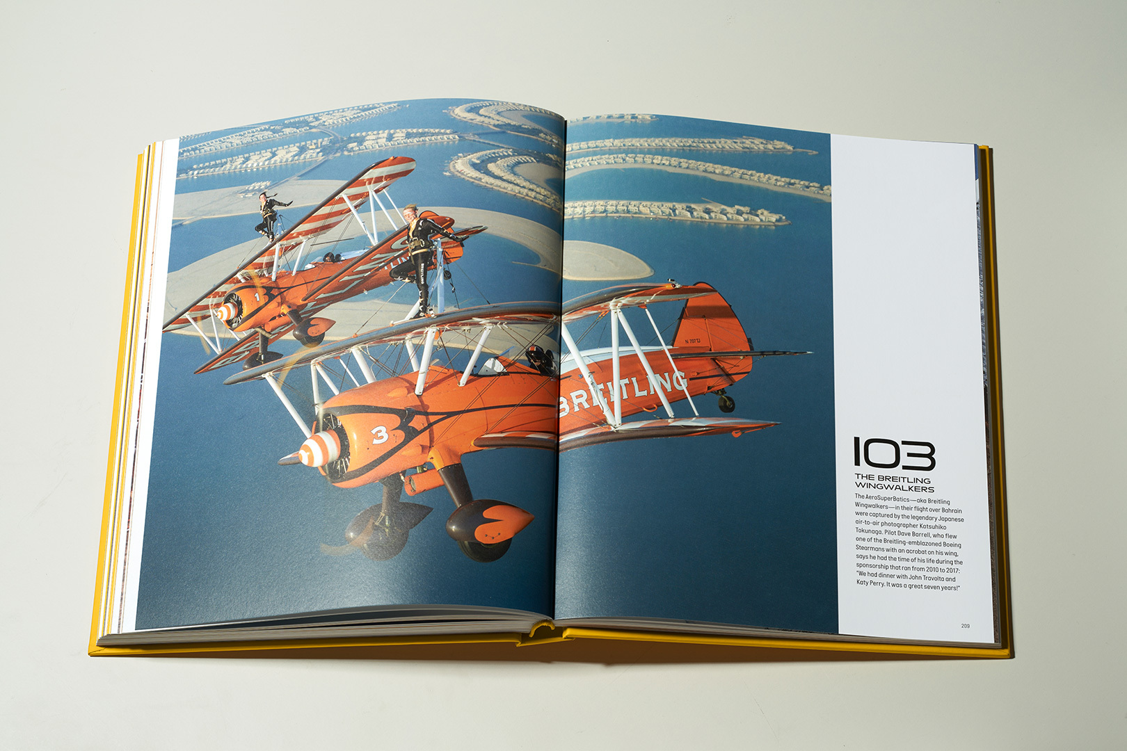 {generated} Livro comemorativo aberto com os Breitling Wingwalkers em biplanos laranja voando sobre uma paisagem costeira