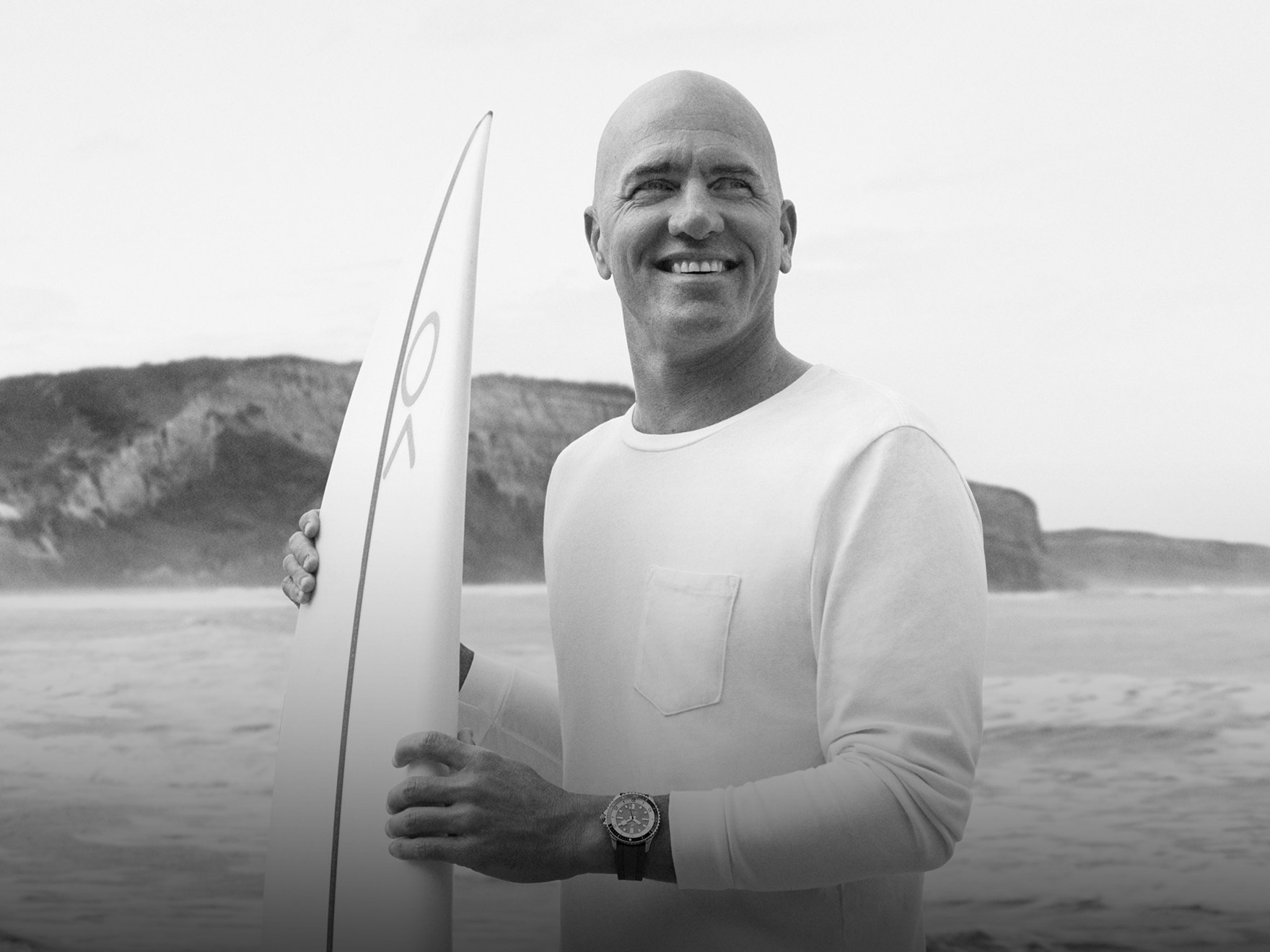 {generated} A surfer holding a board on a rocky shoreline, wearing a Breitling watch that highlights its sporty coastal style