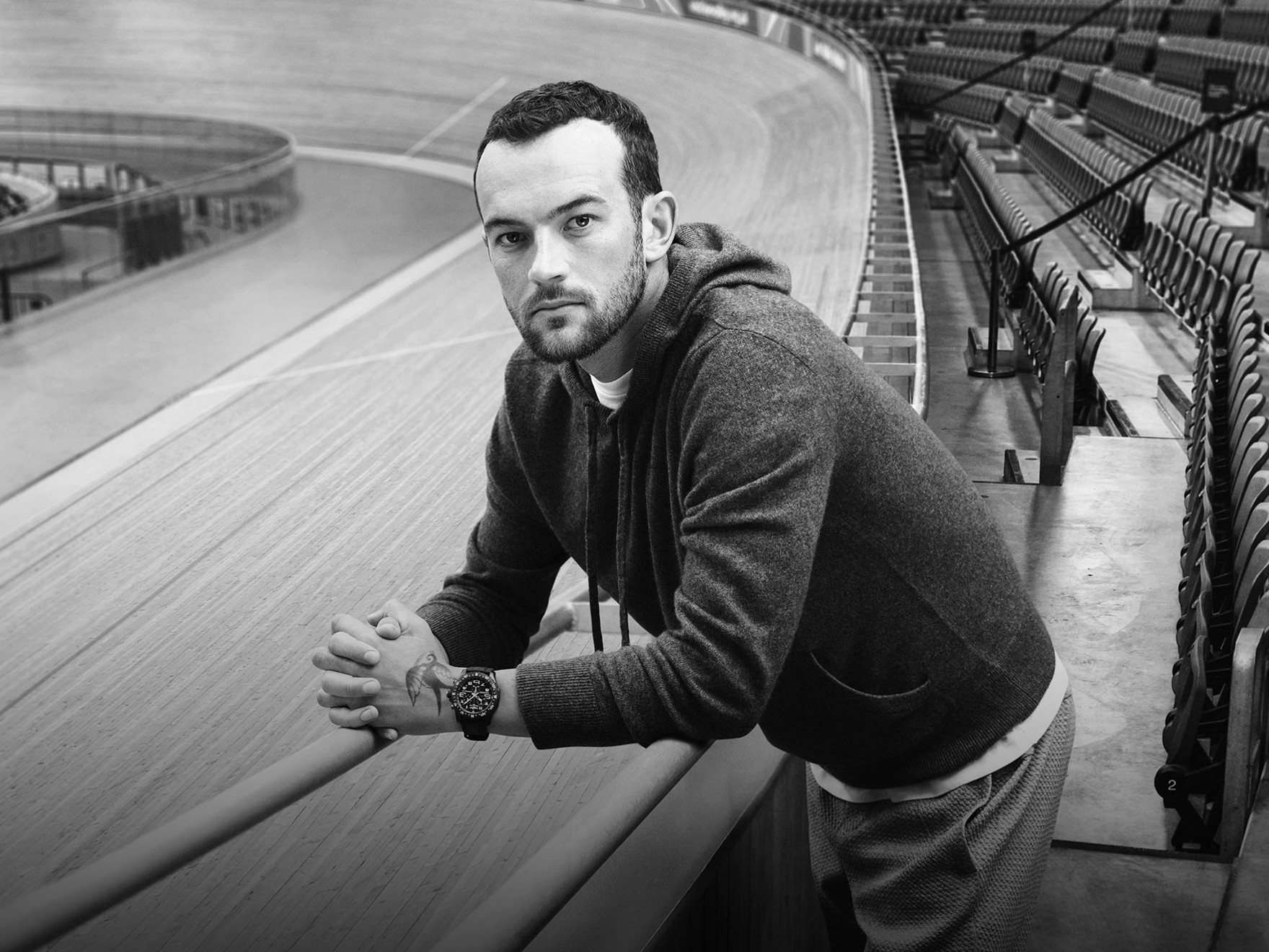 {generated} A man at a velodrome rail wearing a dark hoodie and a bold Breitling watch, shown in a calm athletic setting