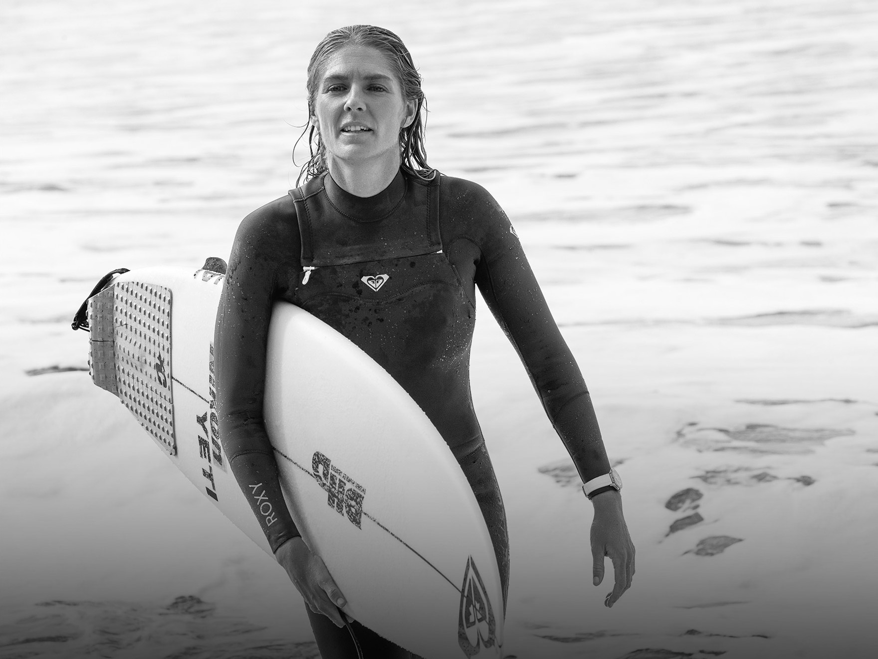 {generated} Female surfer in a wetsuit holding her surfboard at the shoreline, wearing a Breitling watch as waves roll in behind her.
