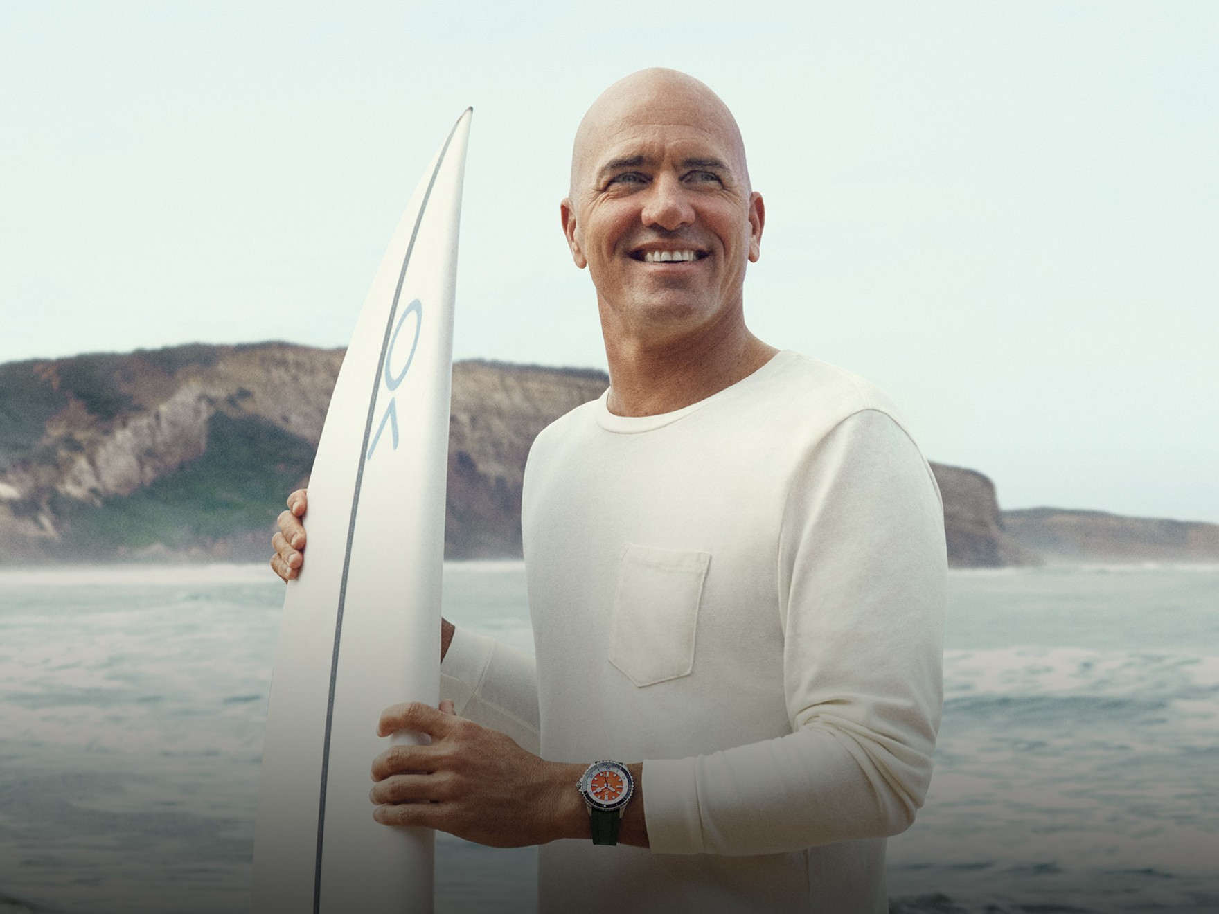 {generated} A surfer holding a board on a rocky shoreline, wearing a Breitling watch that highlights its sporty coastal style