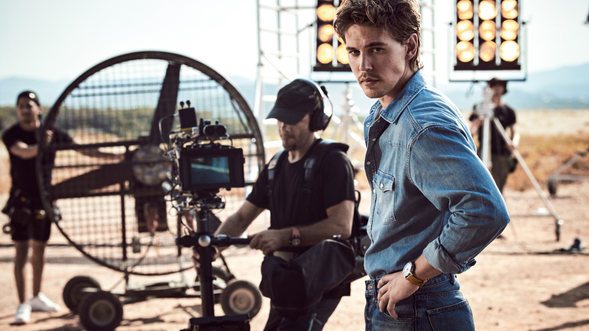 {generated} 日差しの強い撮影現場で、男性がBreitling Top Time B31を着用し、スタッフや機材、照明のそばに立っている。
