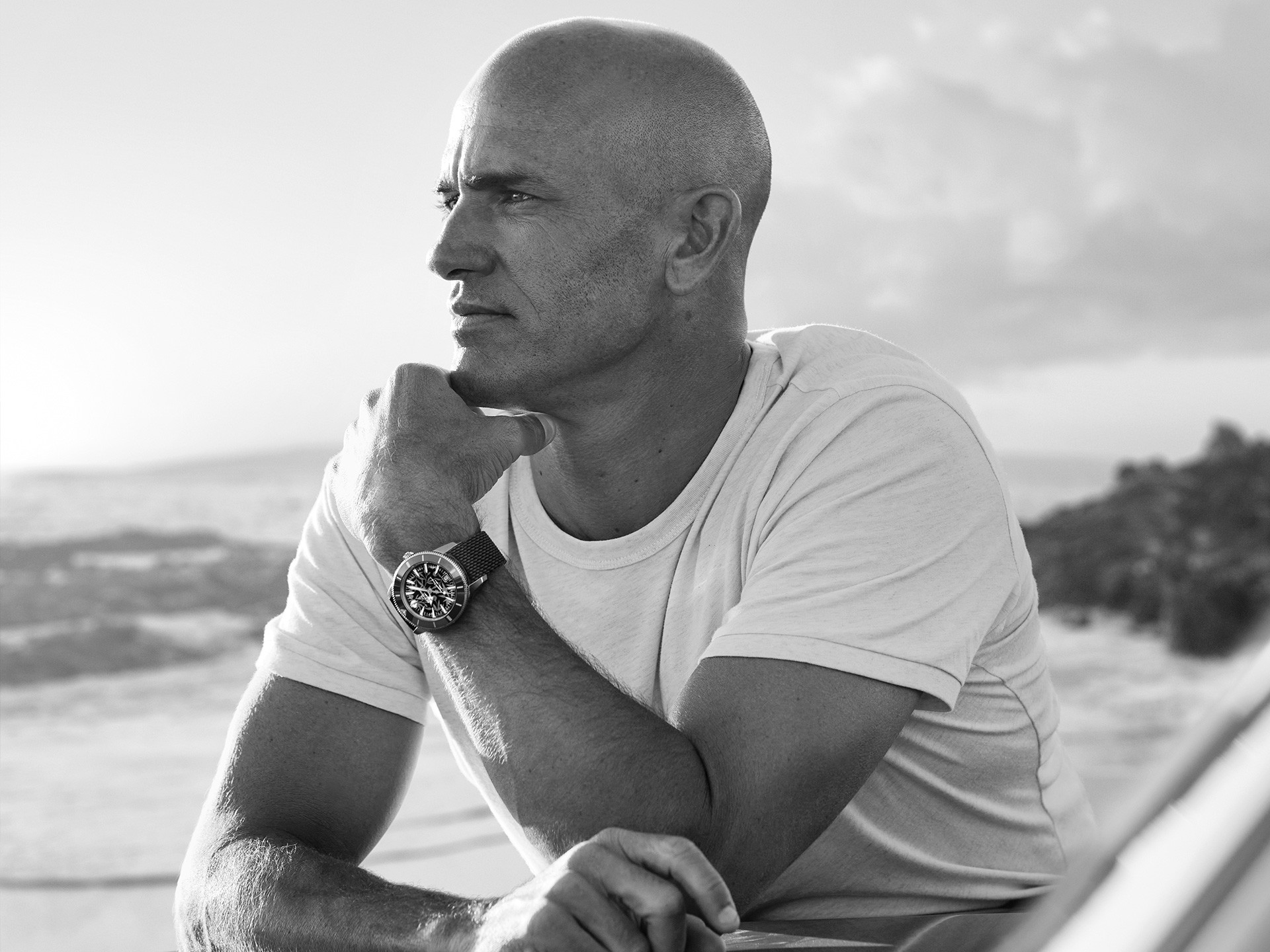 {generated} A surfer holding a board on a rocky shoreline, wearing a Breitling watch that highlights its sporty coastal style