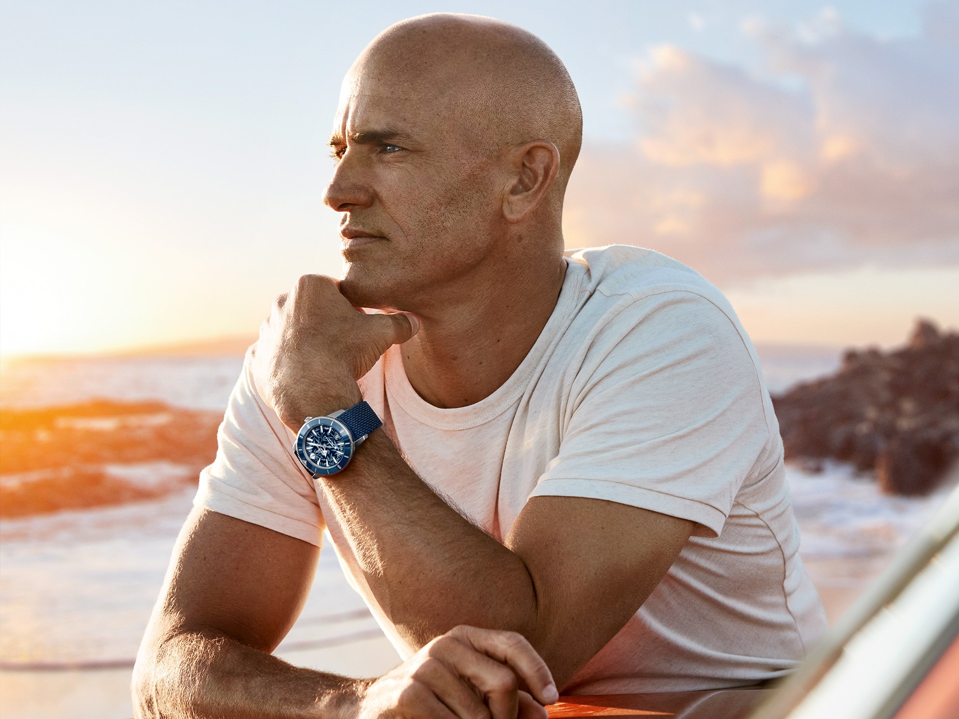 {generated} A surfer holding a board on a rocky shoreline, wearing a Breitling watch that highlights its sporty coastal style