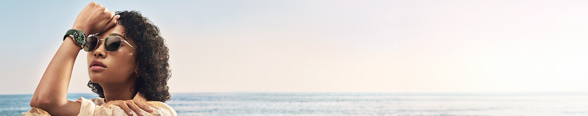 {generated} A person by the sea raises an arm, showcasing a green‑strap Breitling watch against a calm horizon in soft sunlight.