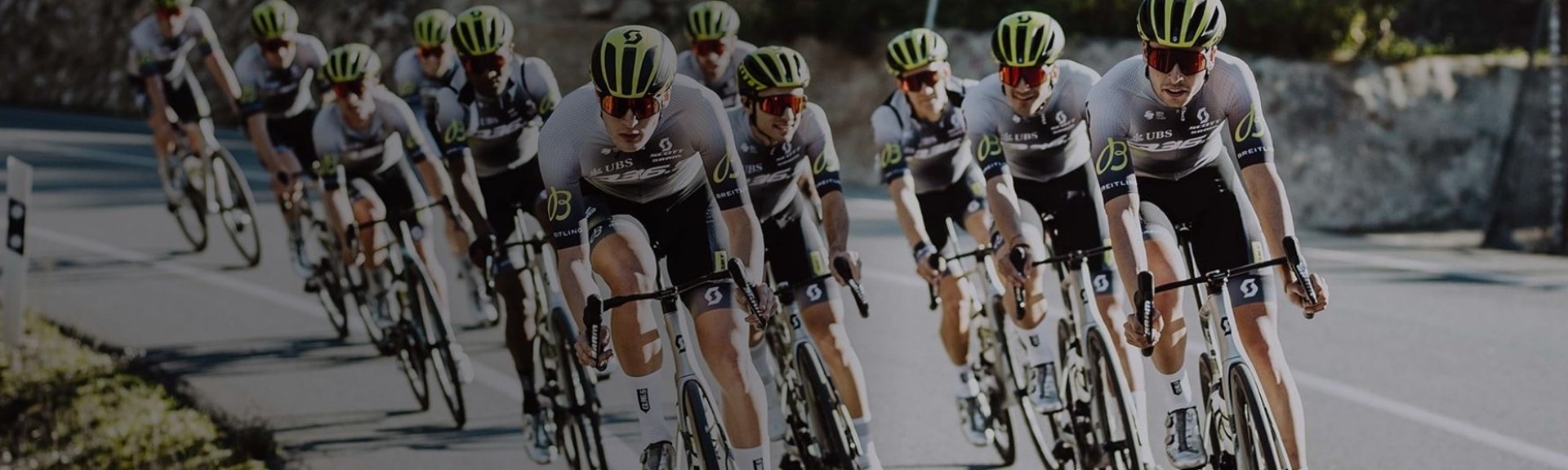 {generated} 身著統一隊服並帶有Breitling標識的車隊在陽光照耀的彎道路段整齊前進。