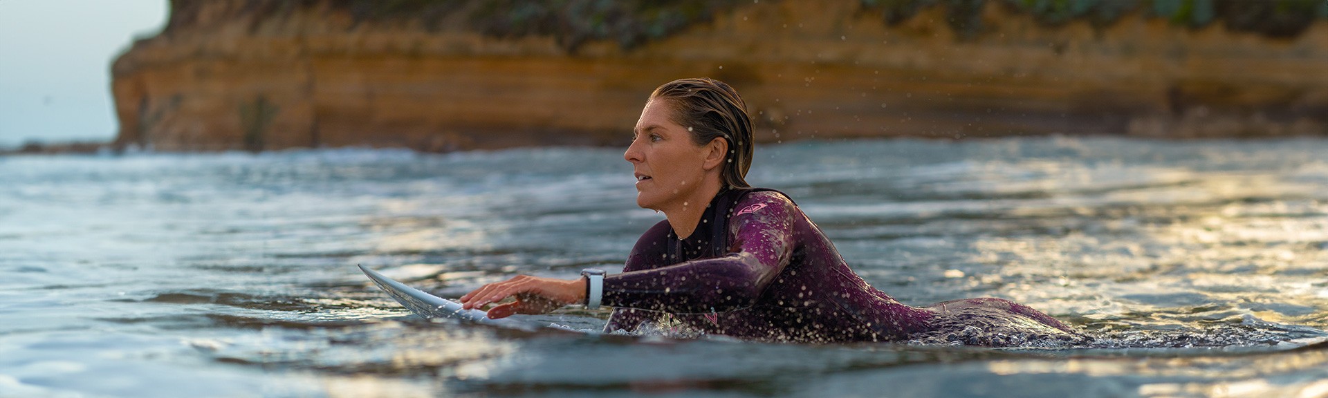 {generated} A woman paddling a surfboard at sunset, wearing a wetsuit and a Breitling watch, with coastal cliffs in the background.