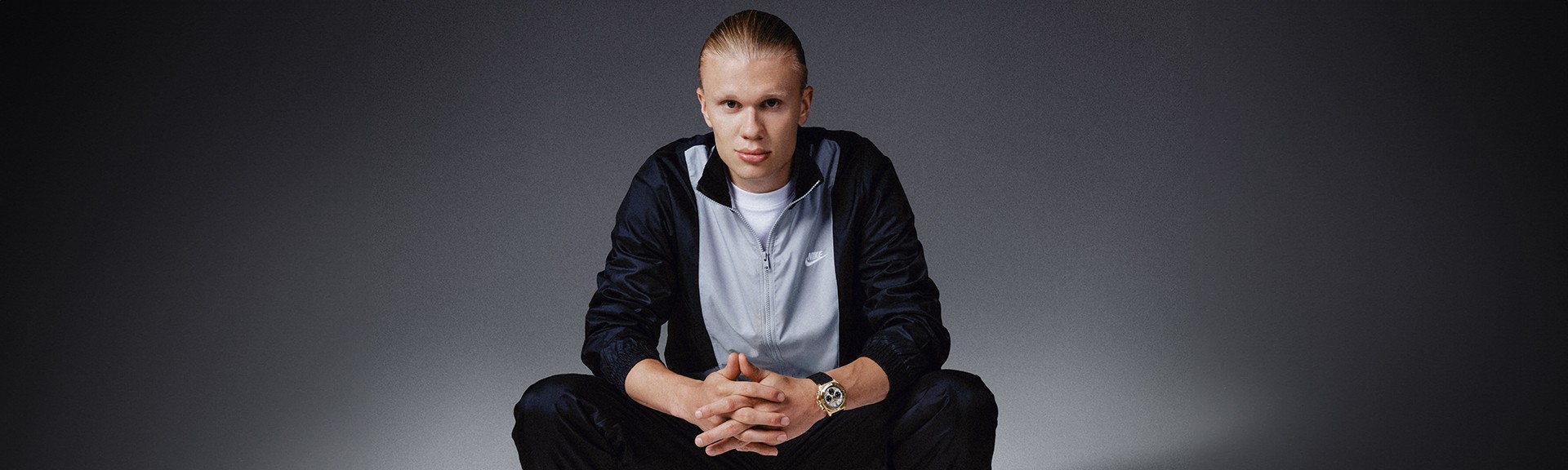 {generated} A seated male ambassador in sportswear highlights a gold Breitling watch against a dark backdrop with a composed posture.
