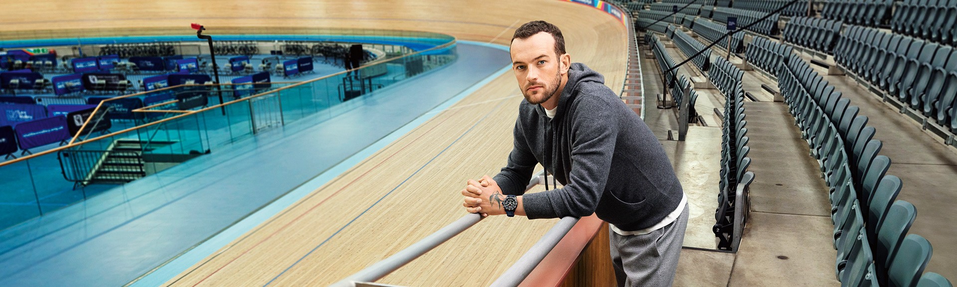 {generated} Man leaning on a velodrome railing wearing a Breitling sports watch with blue strap beside empty tiered seating.