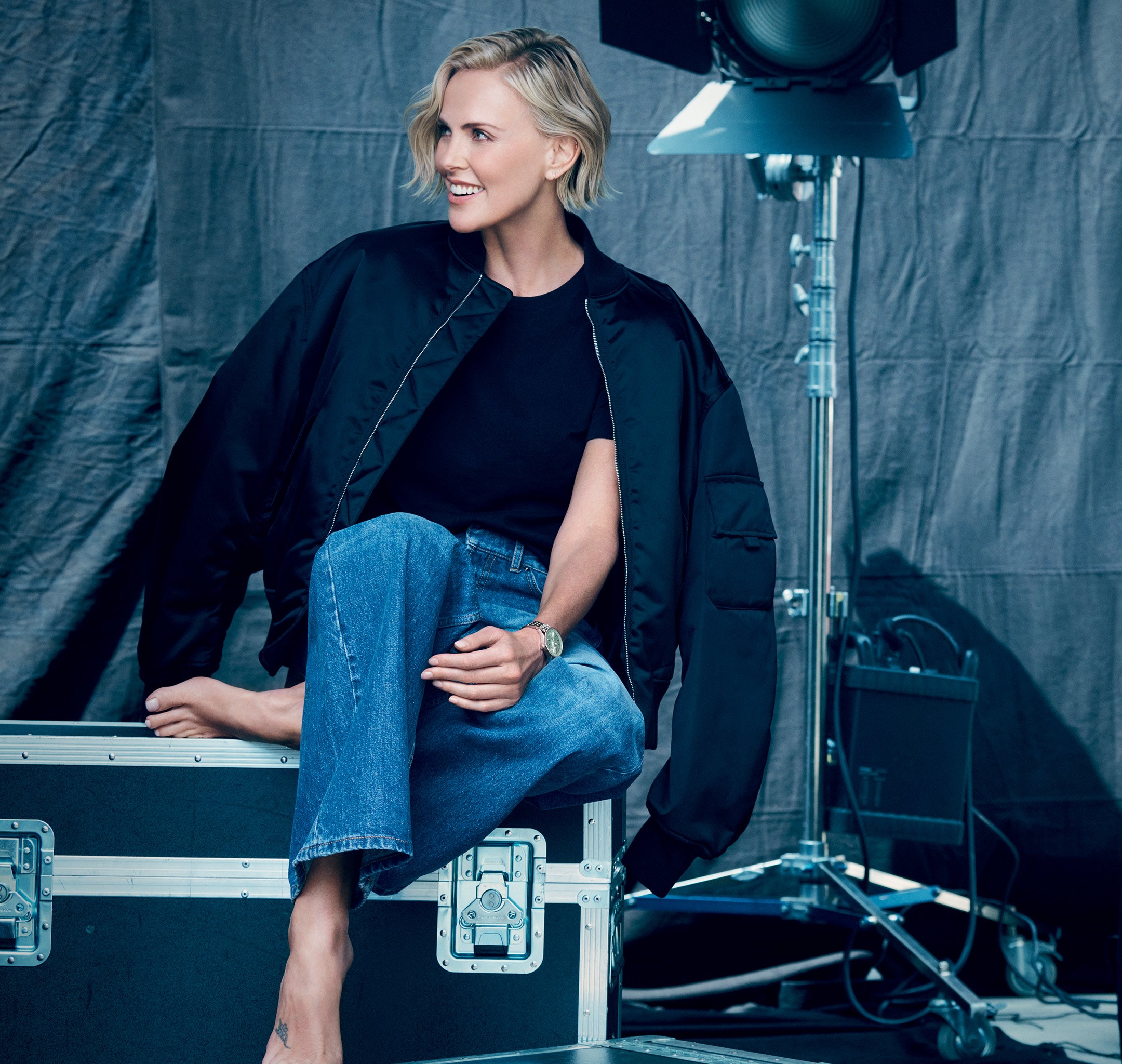 {generated} 身穿牛仔與深色外套的女子赤腳坐在器材箱上，在燈光下突出展示一款百年靈腕錶。
