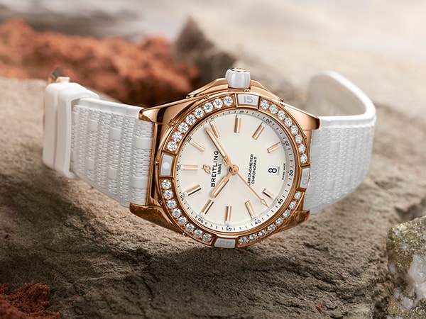 {generated} Montre féminine Breitling dorée à lunette diamantée et bracelet blanc posée sur roche brute sous une lumière douce et chaude.