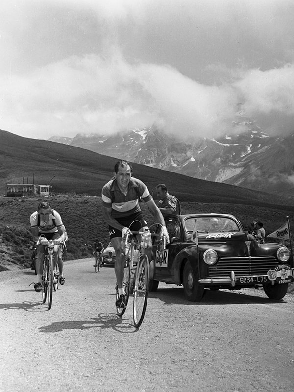 {generated} Bartali lidera a subida montanhosa com ciclistas e um carro de apoio seguindo por uma estrada alpina acidentada.