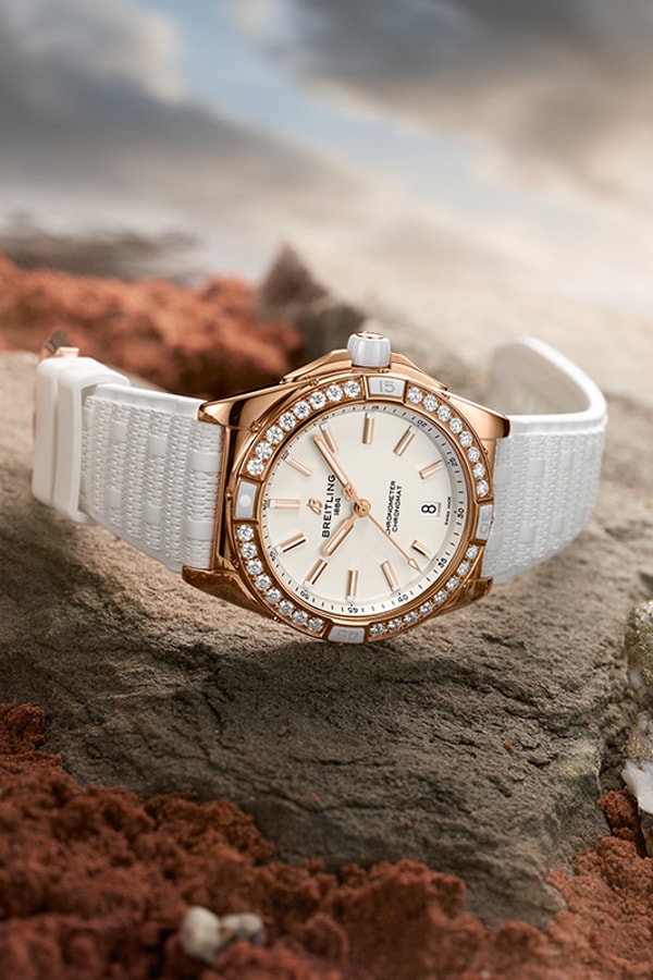 {generated} Montre féminine Breitling dorée à lunette diamantée et bracelet blanc posée sur roche brute sous une lumière douce et chaude.