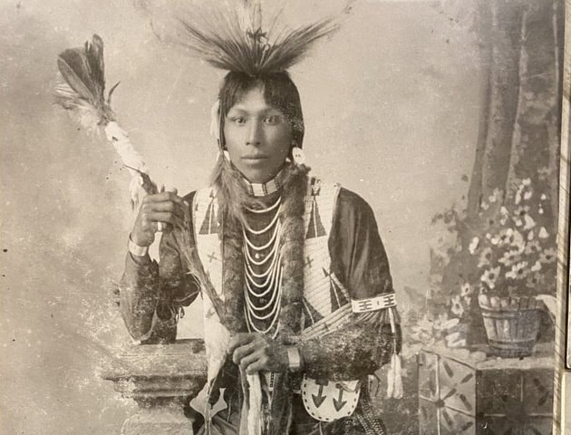 {generated} TU’PAXIN stands in traditional attire holding a decorated staff against a studio backdrop with cultural details.