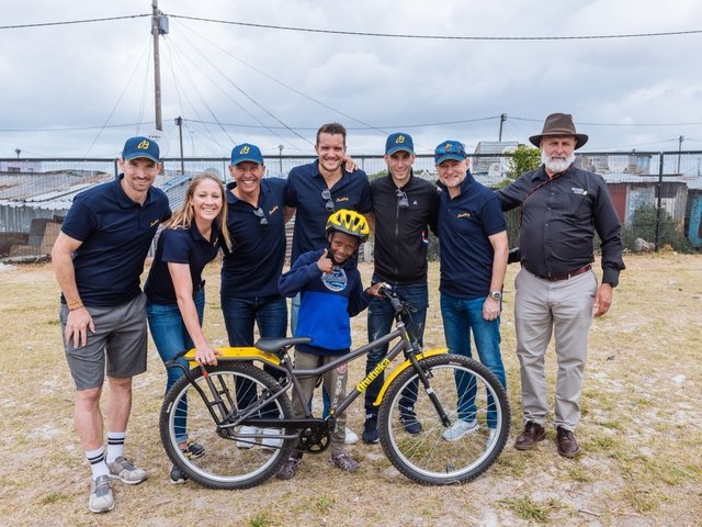 {generated} Équipe Breitling réunie avec un enfant et un vélo jaune, soulignant le soutien communautaire et l’accès à la mobilité