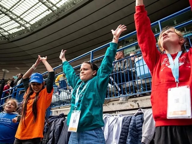 {generated} Des enfants lors d’un événement Premiers de Cordée au stade lèvent les bras avec enthousiasme, entourés de spectateurs dans les tribunes.