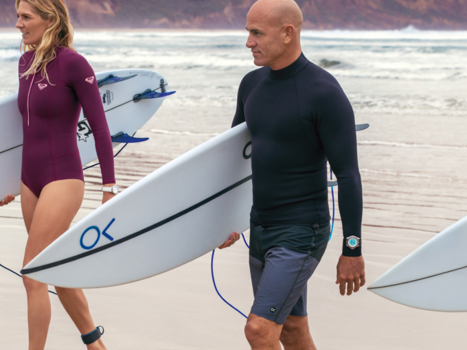 {generated} Surfers walk along a windy beach, with the man wearing a Breitling dive watch emphasising its helium escape valve design