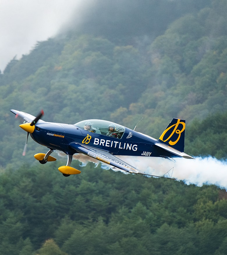 {generated} Breitlingロゴを配した曲技飛行機が煙の軌跡を描き森林背景を背に飛行する様子を捉えた動的なシーン